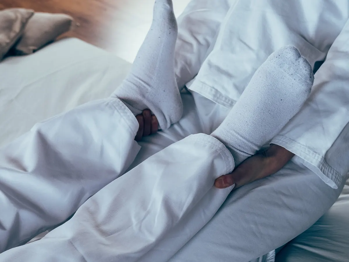 Primo piano: le mani di Cecilia sostengono da sotto le gambe, in calzini bianchi, di una persona distesa sul futon.