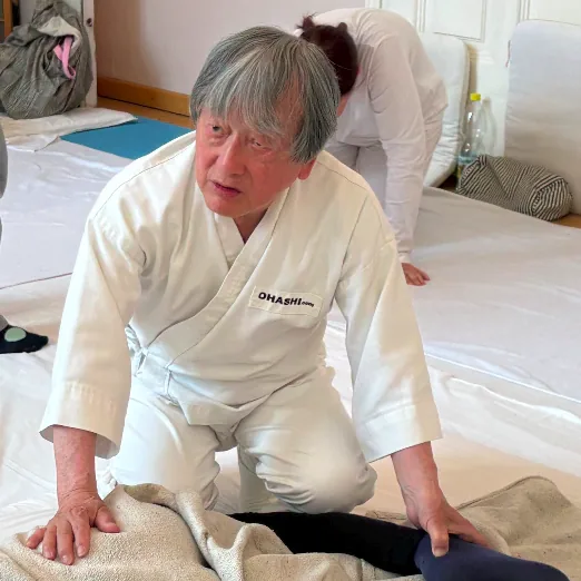 Il maestro Wataru Ohashi, in giacca bianca, inginocchiato su un futon con le mani appoggiate su una persona coperta da una coperta di lino.