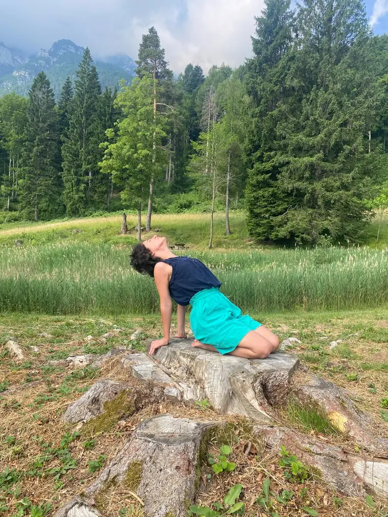 Cecilia seduta su un ceppo, mani appoggiate dietro e torace aperto verso il cielo, in un prato di montagna.