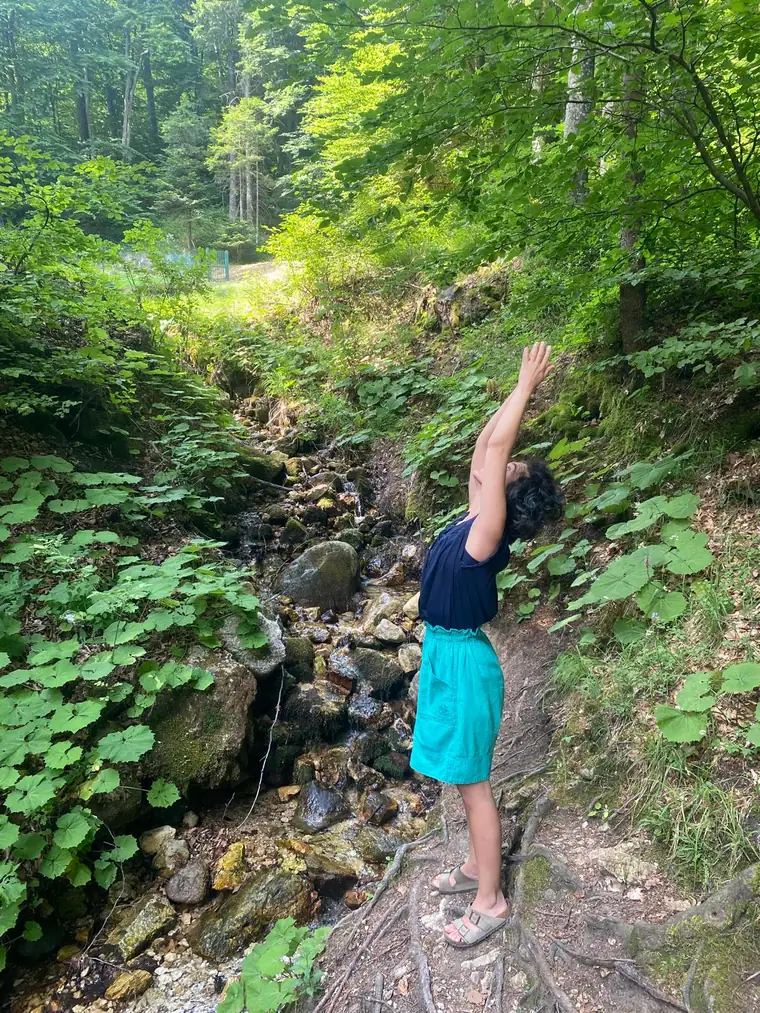 Cecilia in piedi accanto a un torrente nel bosco, un braccio teso verso l'alto tra la vegetazione.