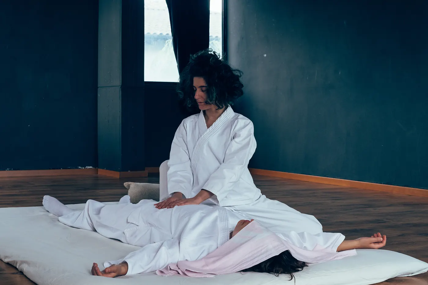 Cecilia, seduta a terra in kimono bianco, appoggia le mani sul fianco di una persona distesa sul futon.