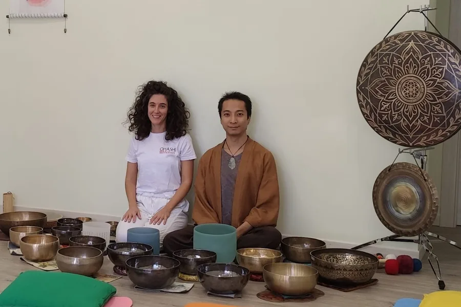 Cecilia e Yip Ngan seduti a terra, sorridenti, davanti a una parete chiara, circondati da campane tibetane e di cristallo; a destra due gong.
